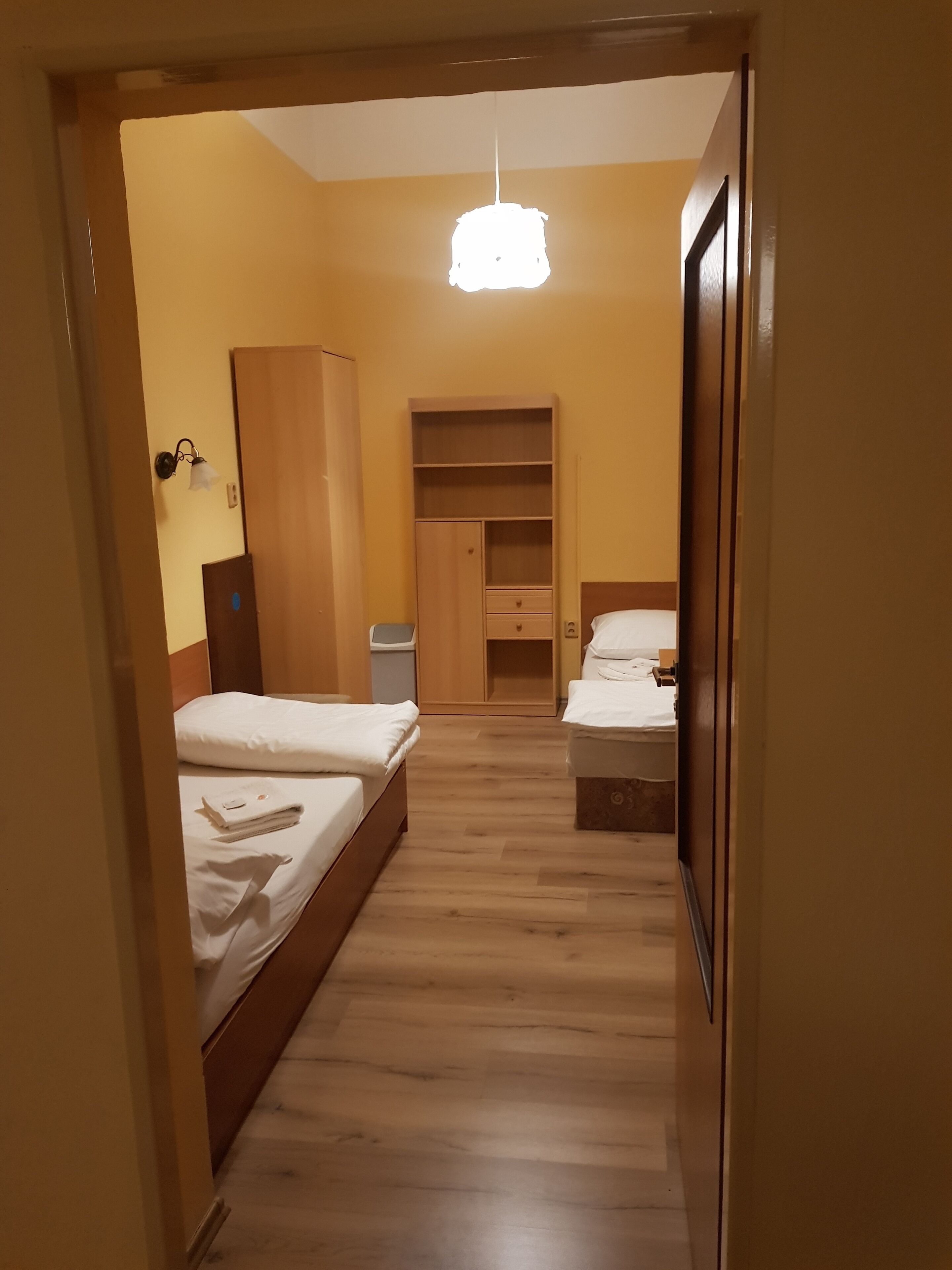 A hotel room with two beds, a wardrobe, a desk, and a hanging light fixture.
