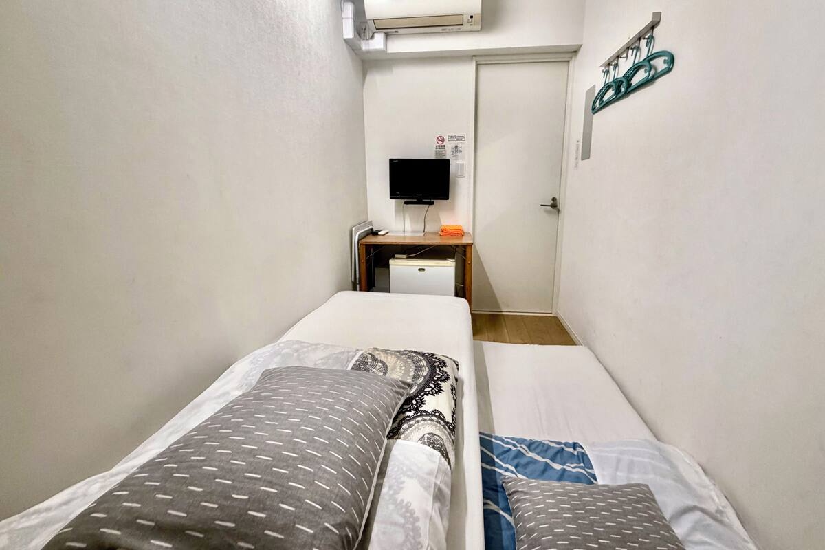 A narrow hotel room with two single beds, a desk with a computer, a flat-screen TV, and an air conditioning unit on the wall.