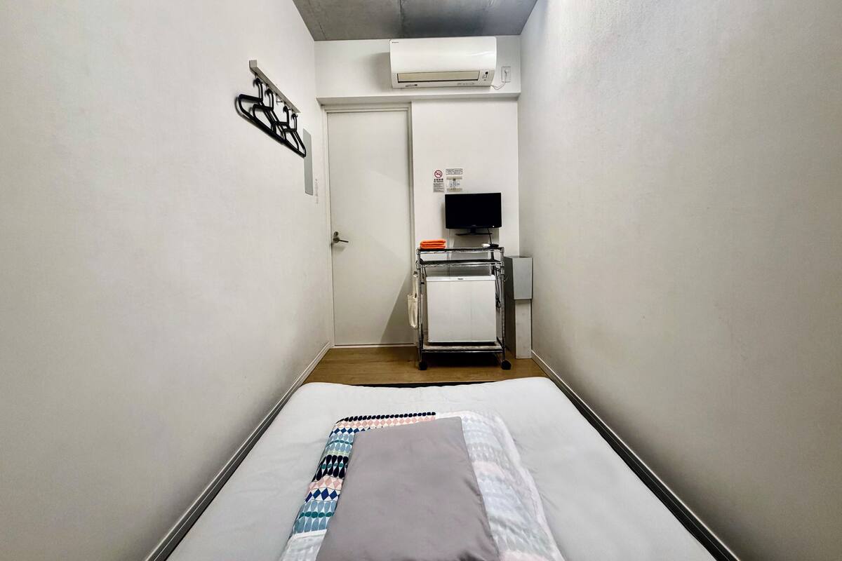 A narrow hotel room with a bed, an air conditioner, a television on a stand, and a door with a sign.