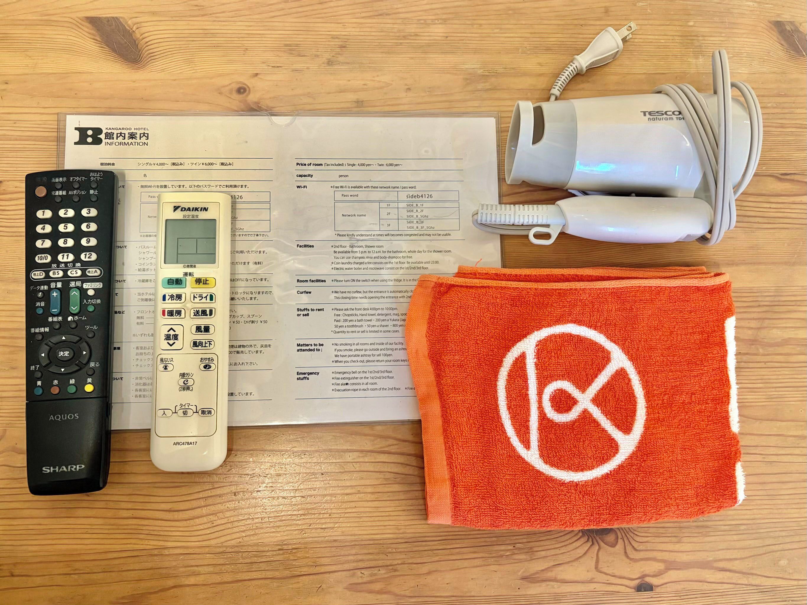 A remote control, a hairdryer, a towel with a logo, and a hotel information card on a wooden surface.