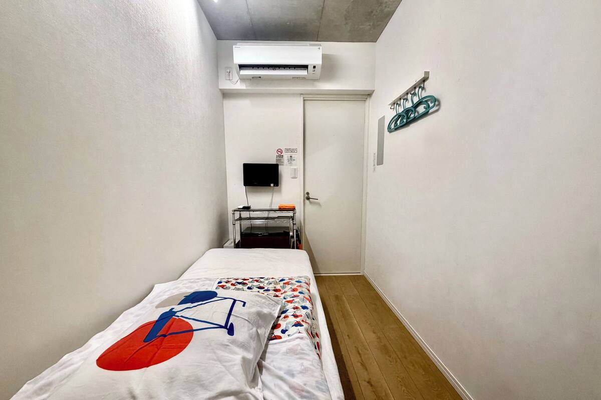 A narrow hotel room with a bed, a desk, an air conditioner, a TV, and a wall-mounted rack.