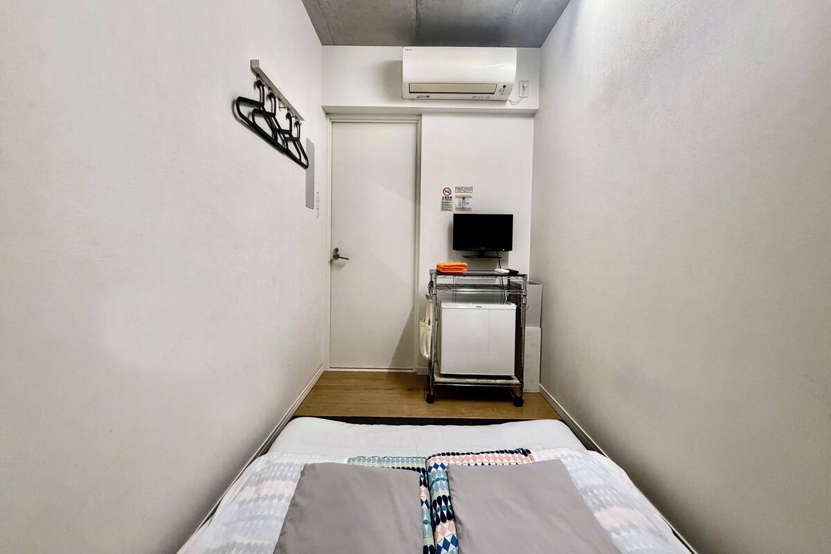 A narrow hotel room with a bed, a television on a stand, an air conditioner, and a door with a sign.