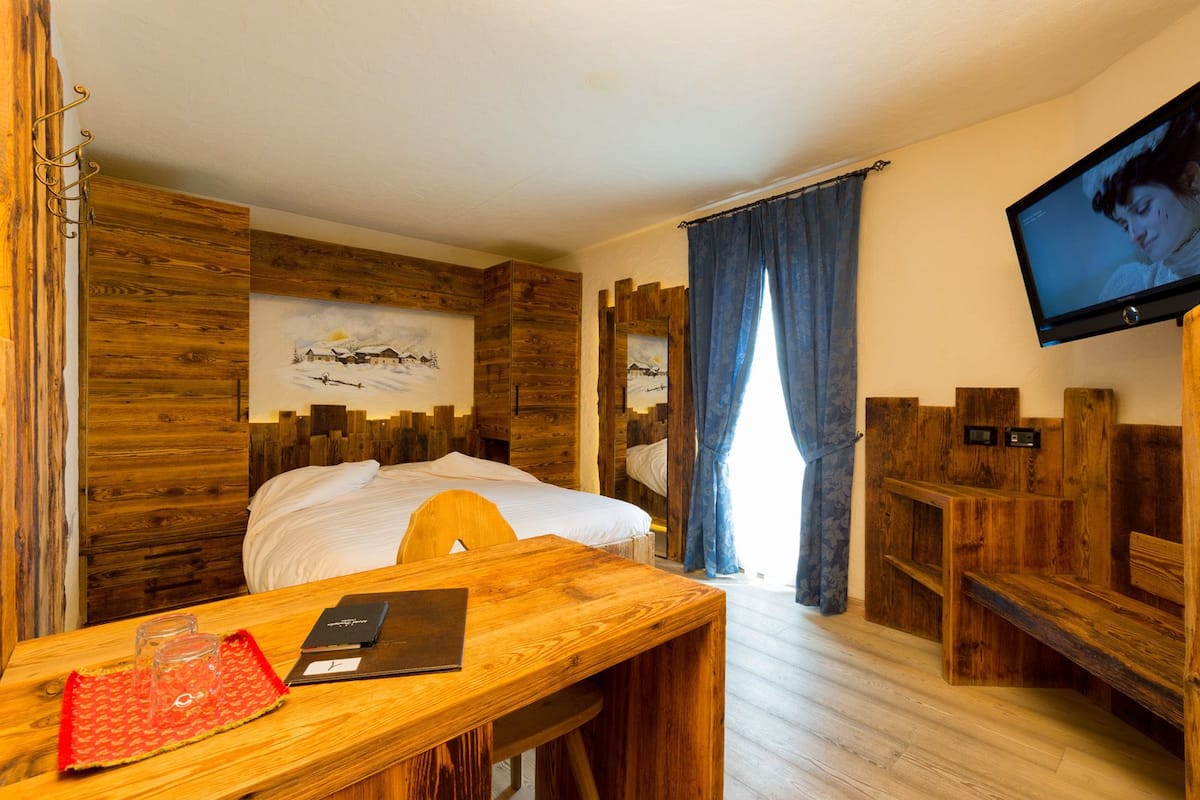 A wooden-themed hotel room with a bed, a desk with a laptop and glasses, a TV, and a painting on the wall.