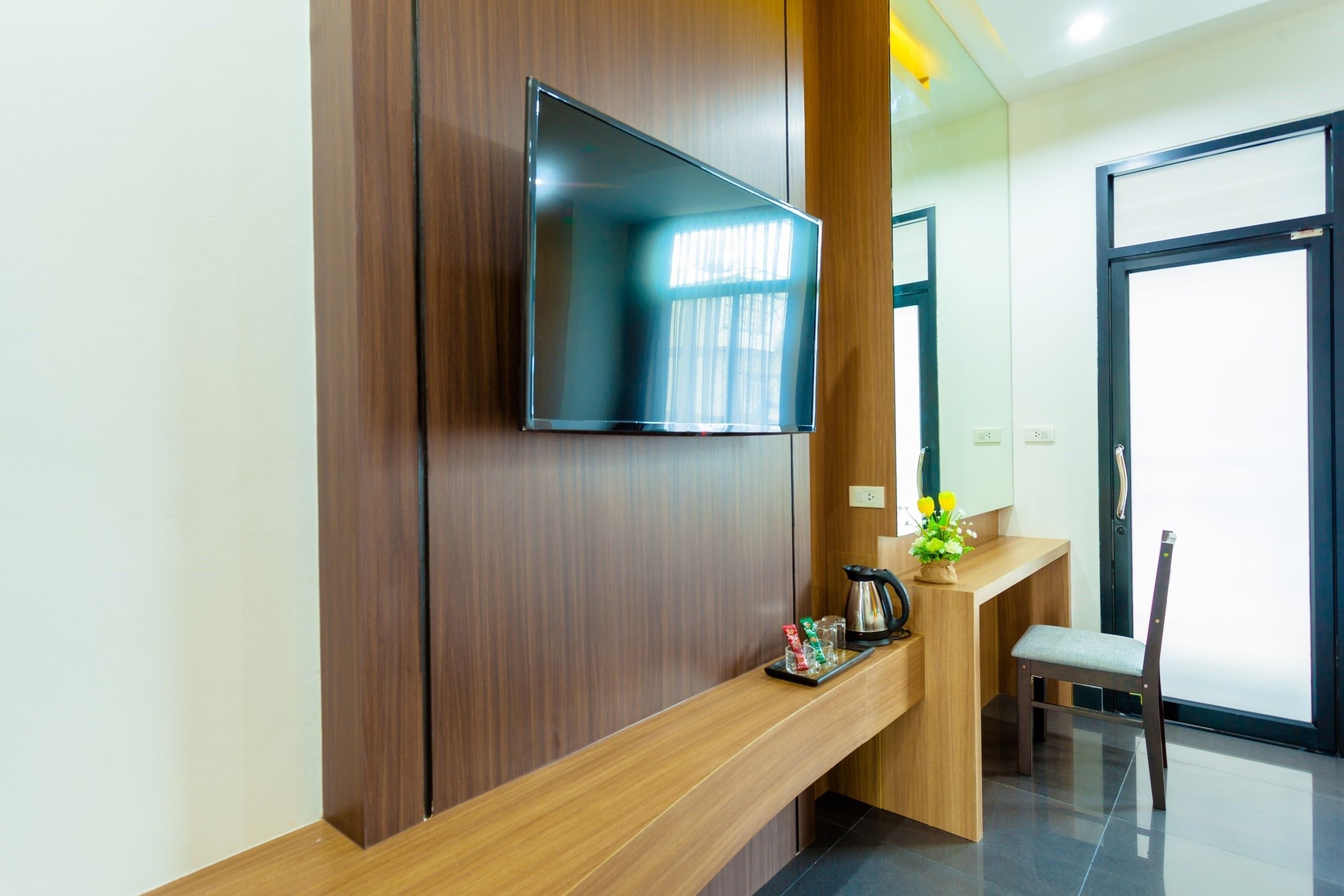 Una moderna zona de recepción de hotel con un televisor de pantalla plana montado sobre un panel de madera, un mostrador de madera con una cafetera, una planta pequeña y una silla.