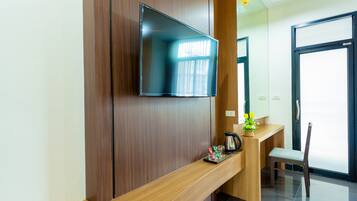 A modern hotel reception area with a flat-screen TV mounted on a wooden panel, a wooden counter with a coffee maker, a small plant, and a chair.