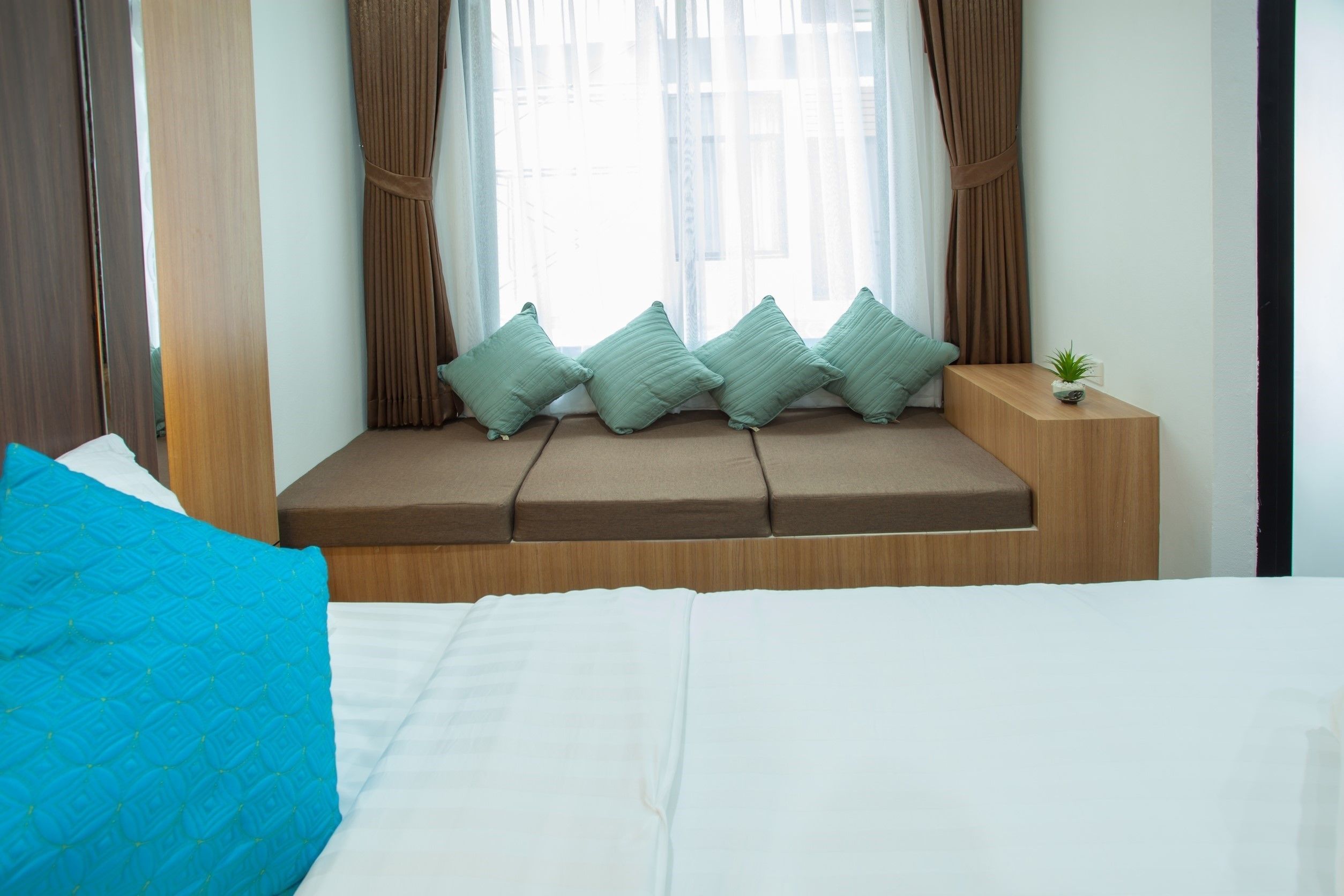 Un dormitorio con cama, un rincón ventana con cojines y una planta pequeña en una mesita de noche.
