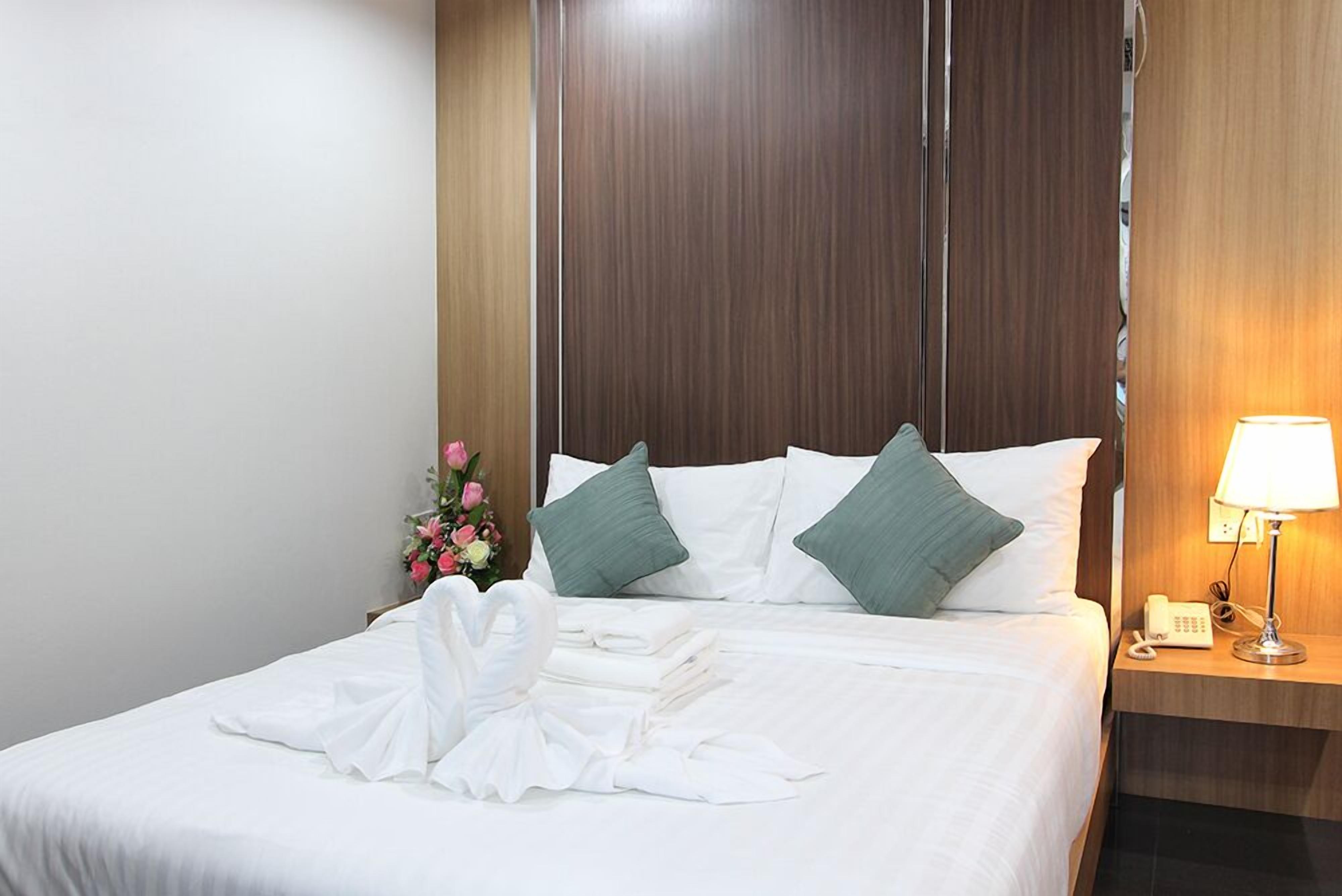 A neatly made bed with white linens and green pillows, a bouquet of flowers, and a folded white towel on the bed.