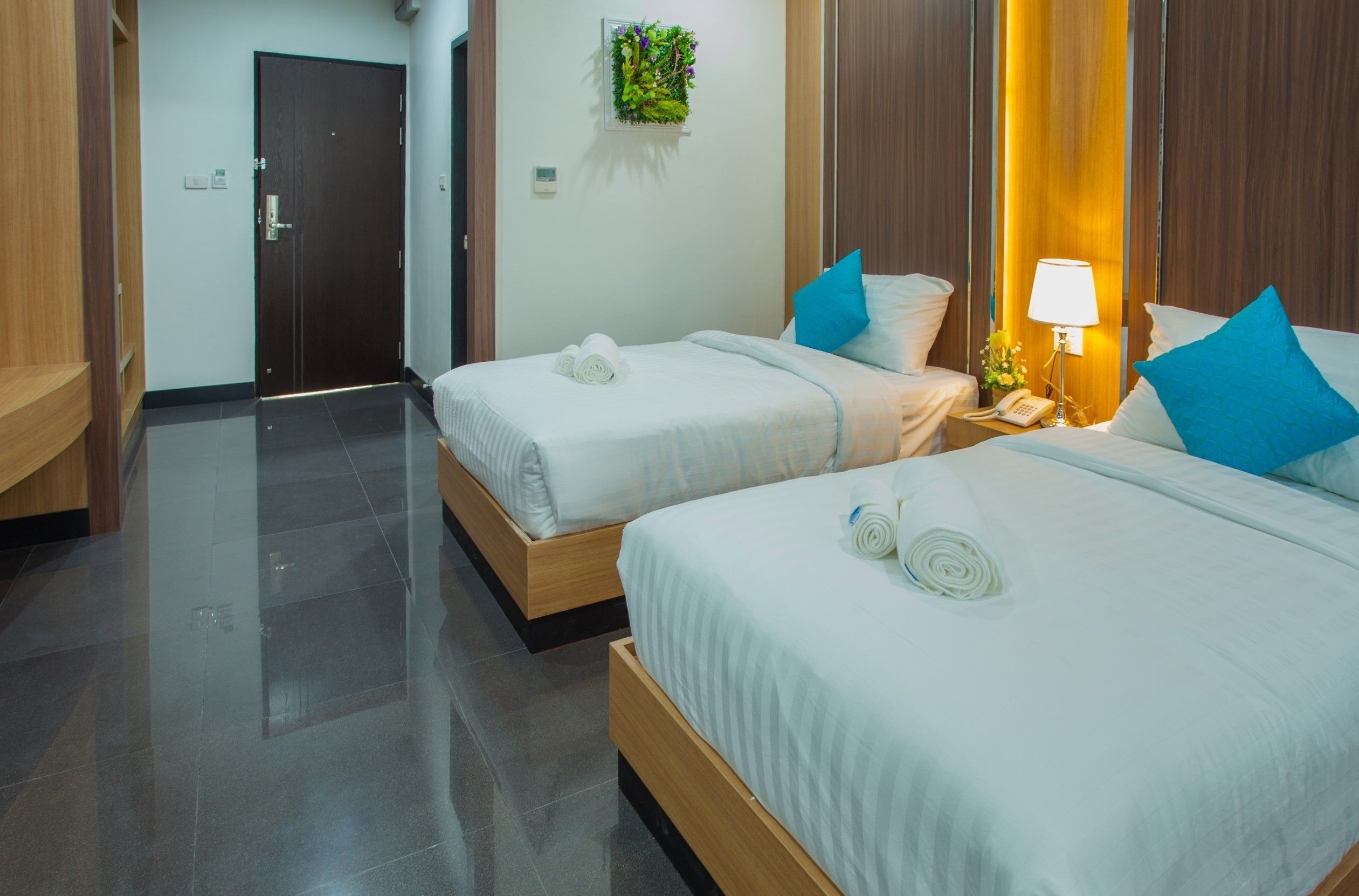 A hotel room with two beds, a bedside table with a lamp, a small plant on the wall, and a door in the background.