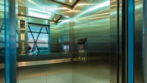 Interior de ascensor con paredes metálicas, puerta de vidrio y panel digital que muestra los pisos.