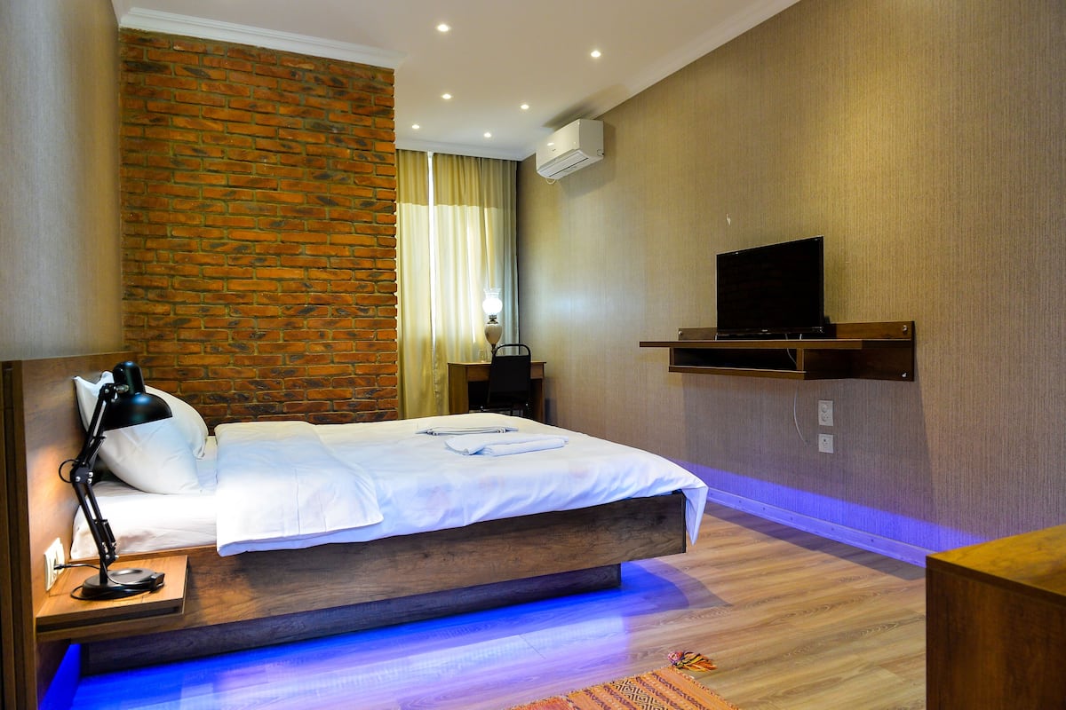 A modern hotel room with a brick accent wall, a flat-screen TV mounted on the wall, a bedside table with a lamp, and a neatly made bed.