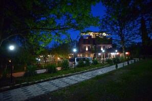 A large, multi-story building with a balcony, surrounded by trees and a paved walkway.