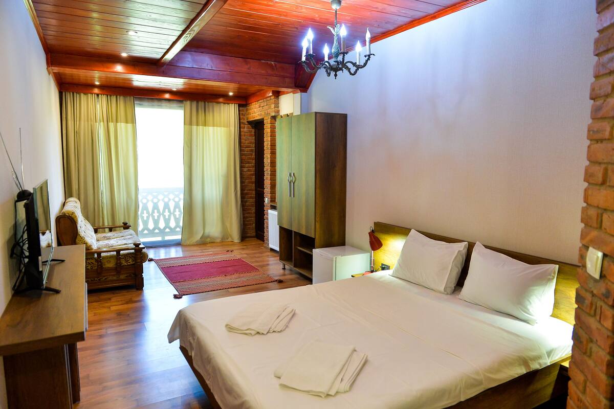 A hotel room with a large bed, a wooden ceiling, a brick wall, and a balcony with curtains.