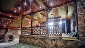 Interior rústico com vigas de madeira aparentes, lareira de tijolos e varanda com grade decorativa.