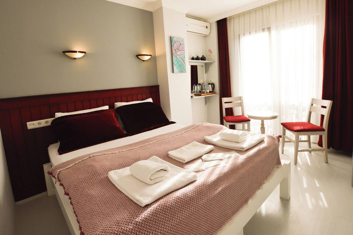 A neatly made bed with white and red linens, a bedside table, a chair, and a window with curtains.