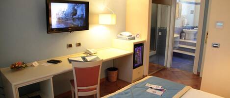 A hotel room with a bed, a desk with a computer, a TV mounted on the wall, and a bathroom visible through an open door.