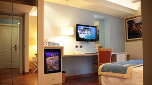 Hotelkamer met een bed, een bureau met een computer en een televisie, een stoel en een tafeltje met een lamp.