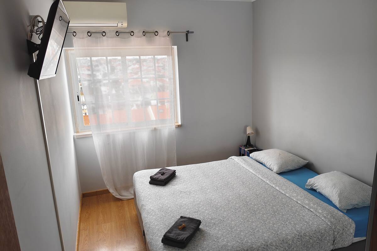 A bedroom with a bed, a nightstand, a lamp, a window with curtains, and an air conditioning unit.