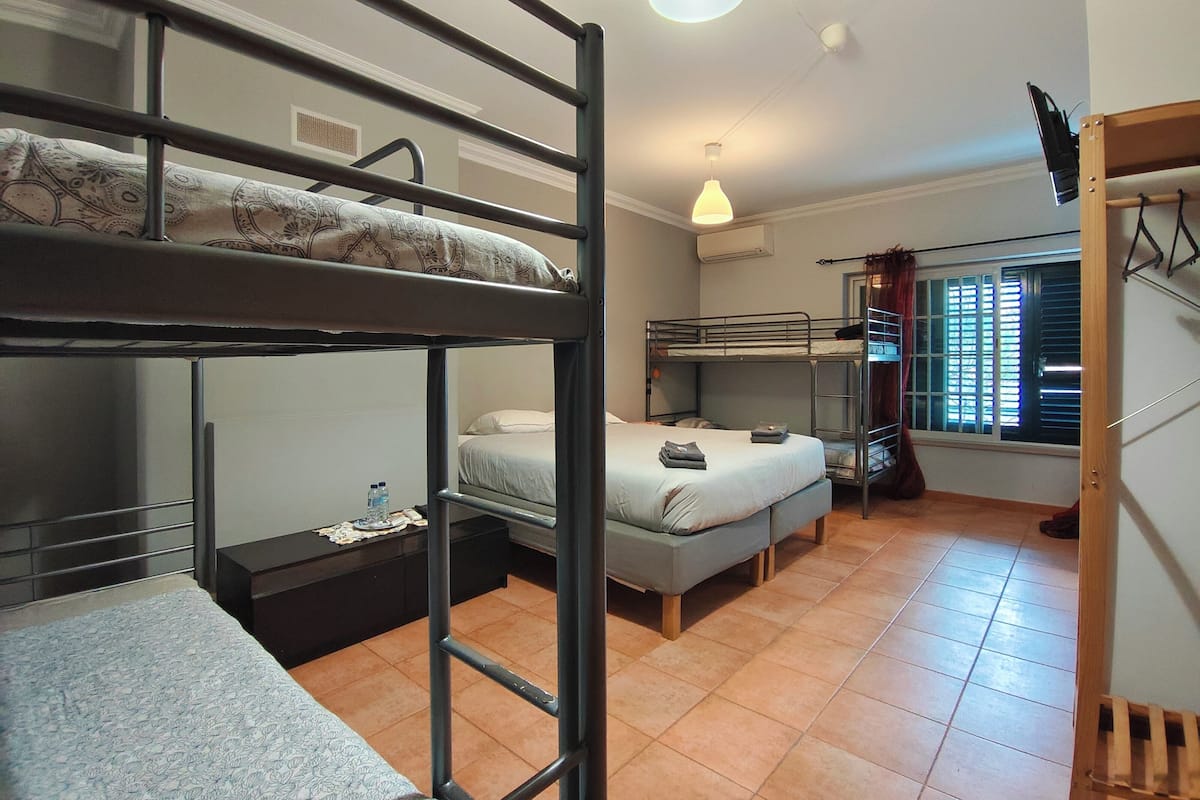 A dormitory room with bunk beds, a desk, a chair, and a window with blinds.