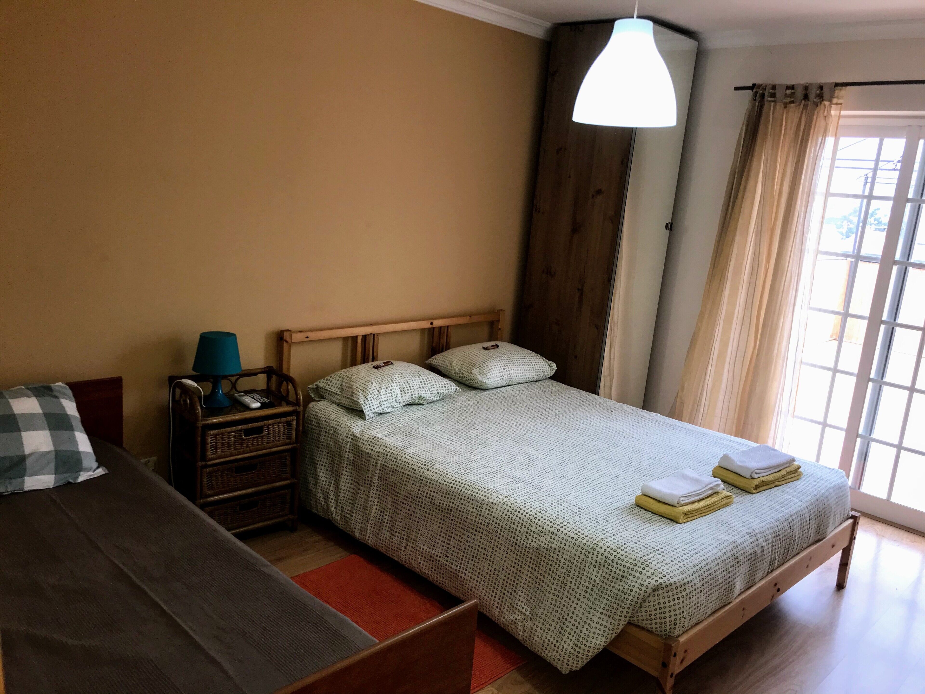 Un dormitorio con cama, mesita de noche, lámpara, armario de madera y ventana con cortinas.
