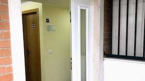 Una habitación con suelo de madera, una puerta blanca que da a un pasillo, una silla de plástico blanco y un bote de basura negro.