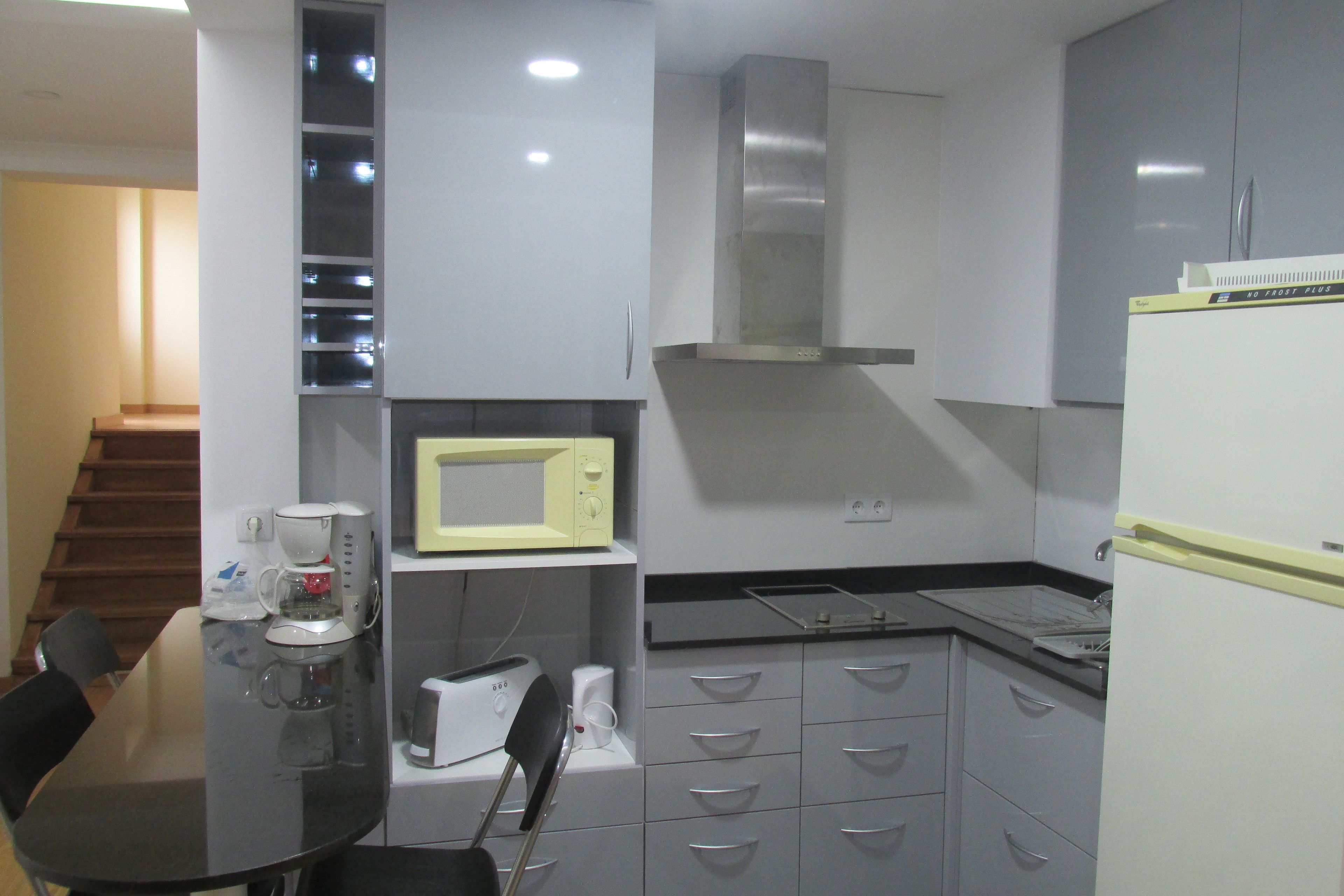 Uma cozinha compacta com micro-ondas, torradeira e cafeteira na bancada, exaustor de aço inoxidável acima e uma geladeira à direita.