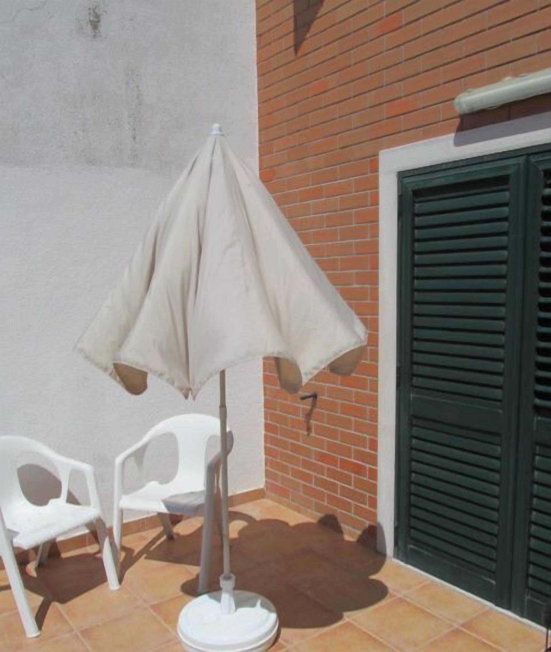Eine Terrasse mit Tisch und Stühlen, einem beigen Sonnenschirm und einer Ziegelwand mit einem Rollo.