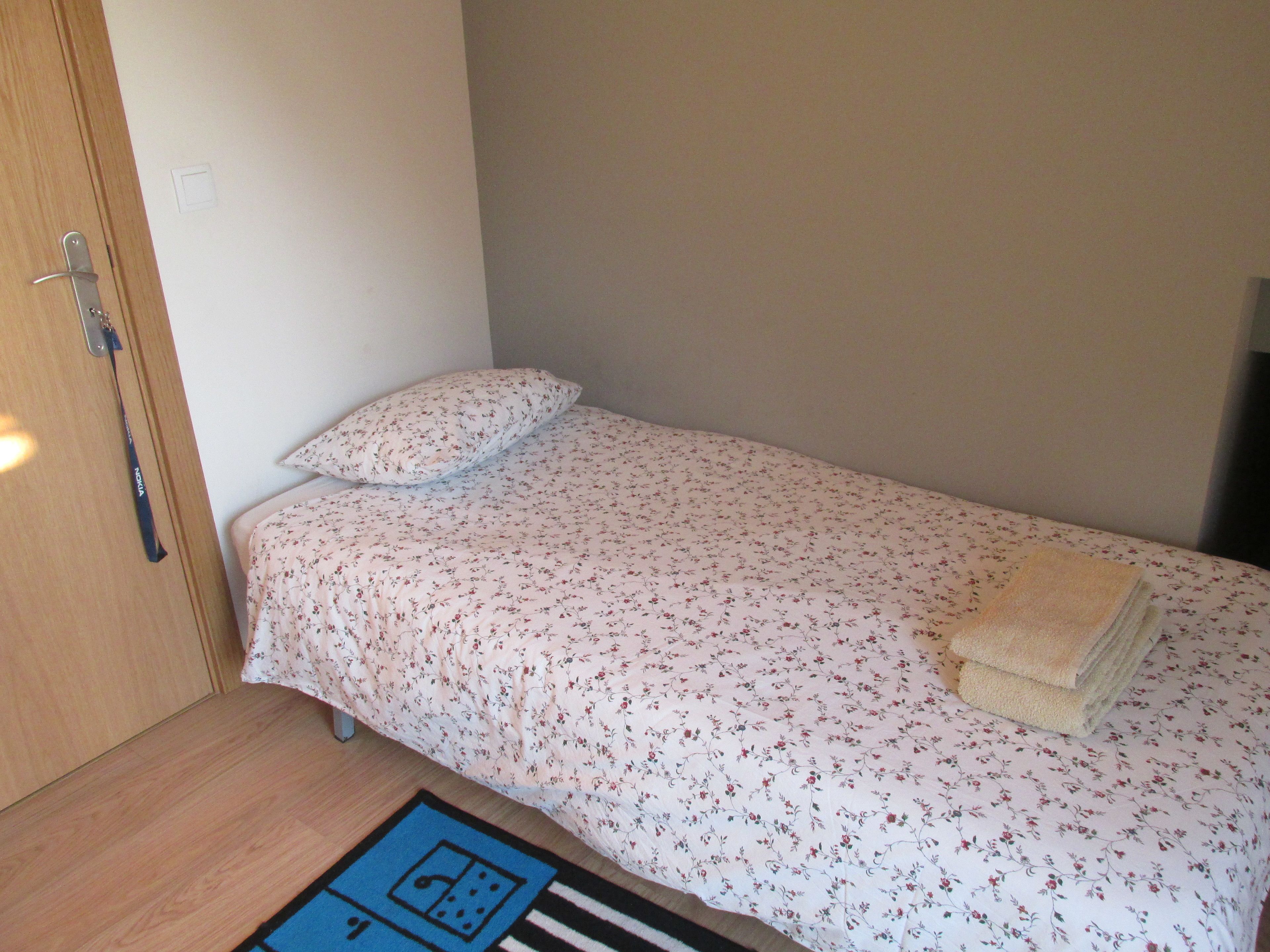 A small, single-bed room with a floral bedspread, a bedside lamp, and a blue rug with a geometric pattern.
