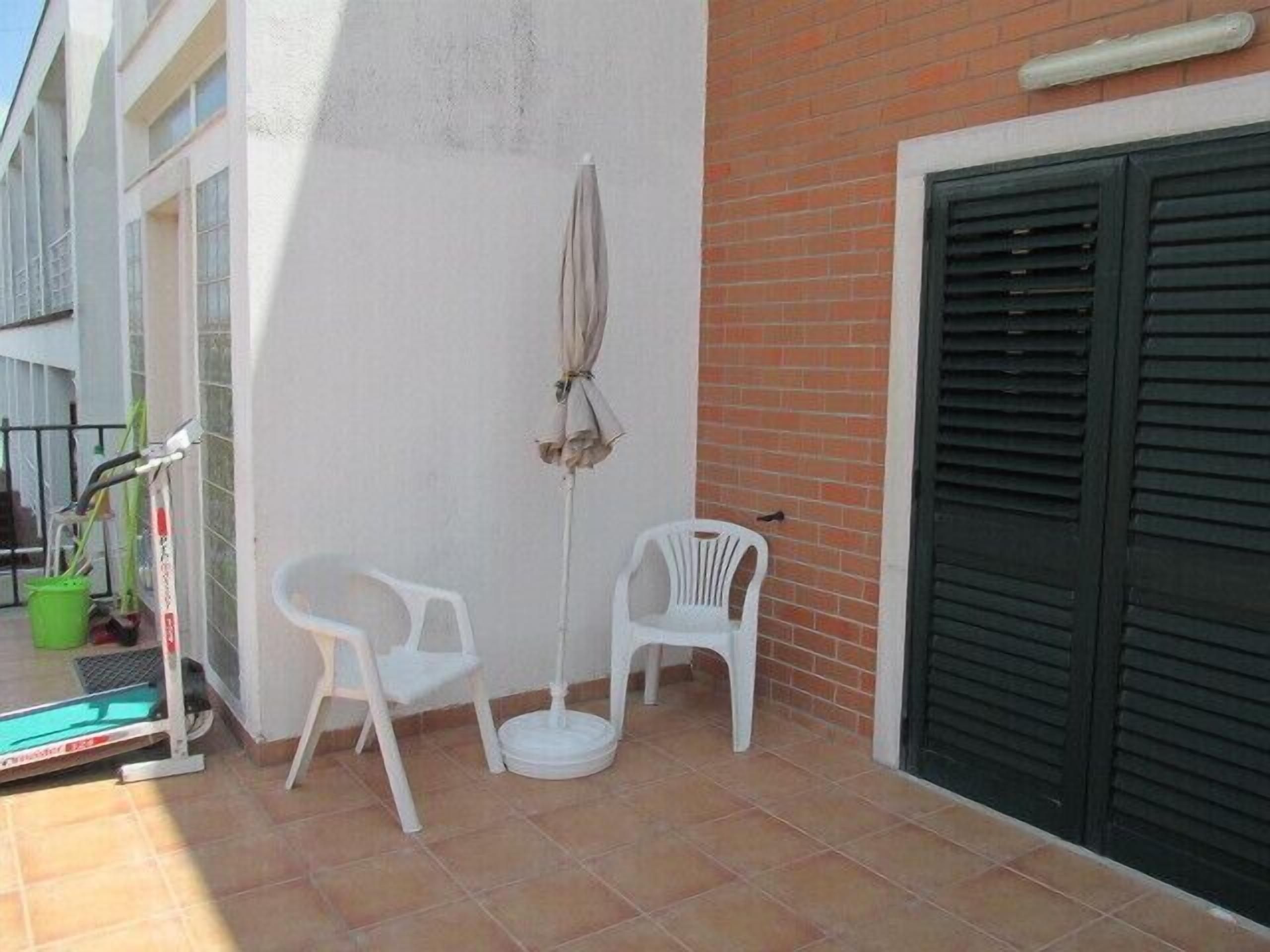 Una pequeña zona de estar al aire libre con dos sillas de plástico blanco, un paraguas, un cubo verde y una herramienta de limpieza en el suelo.