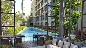 A modern outdoor pool area with a wooden deck, seating, and a view of a high-rise building.