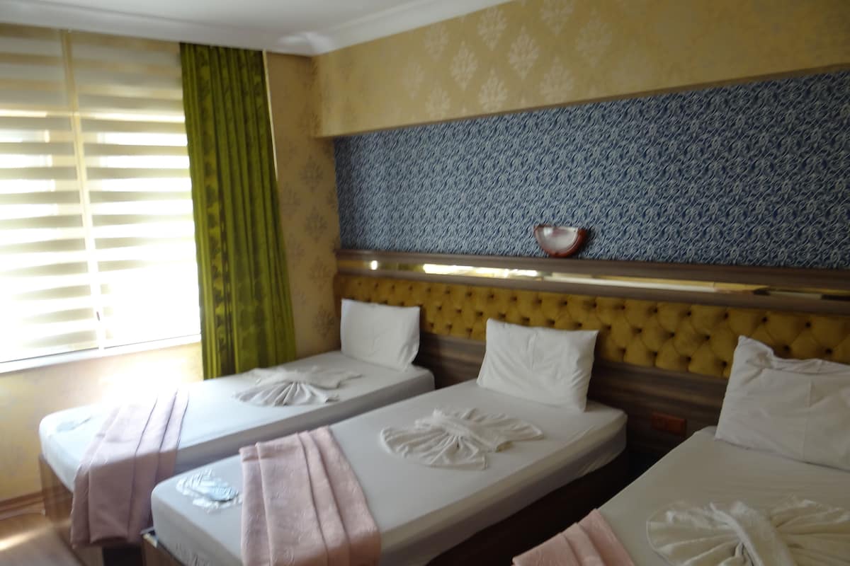 A hotel room with two single beds, each with a neatly folded towel. The room has a patterned wallpaper, a wooden headboard, and a window with blinds.