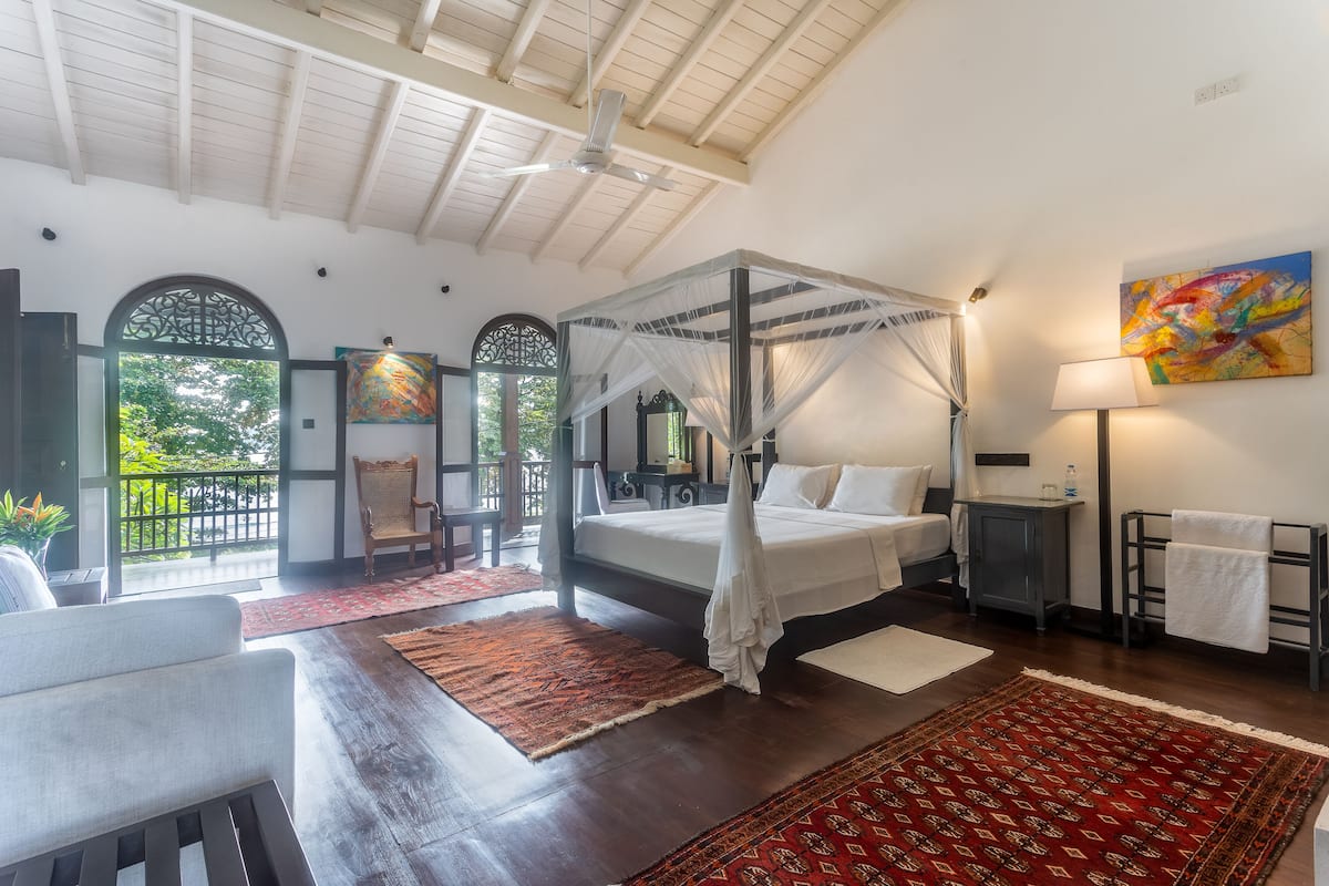 A four-poster bed with a canopy, a red patterned rug, a white sofa, and a wooden floor.
