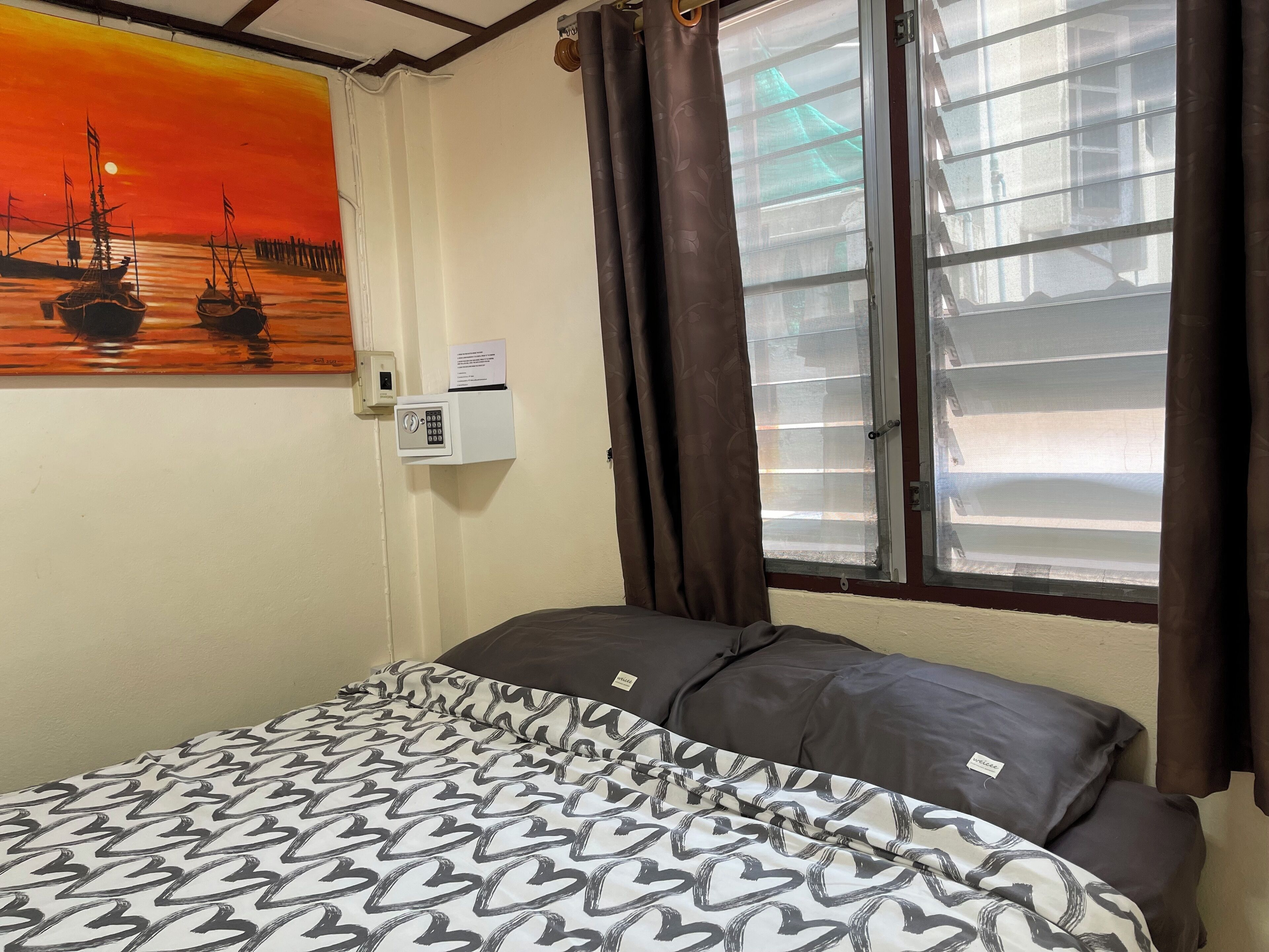 Um quarto com uma cama, um quadro de um pôr do sol sobre a água, uma janela com cortinas e um termostato fixado na parede.