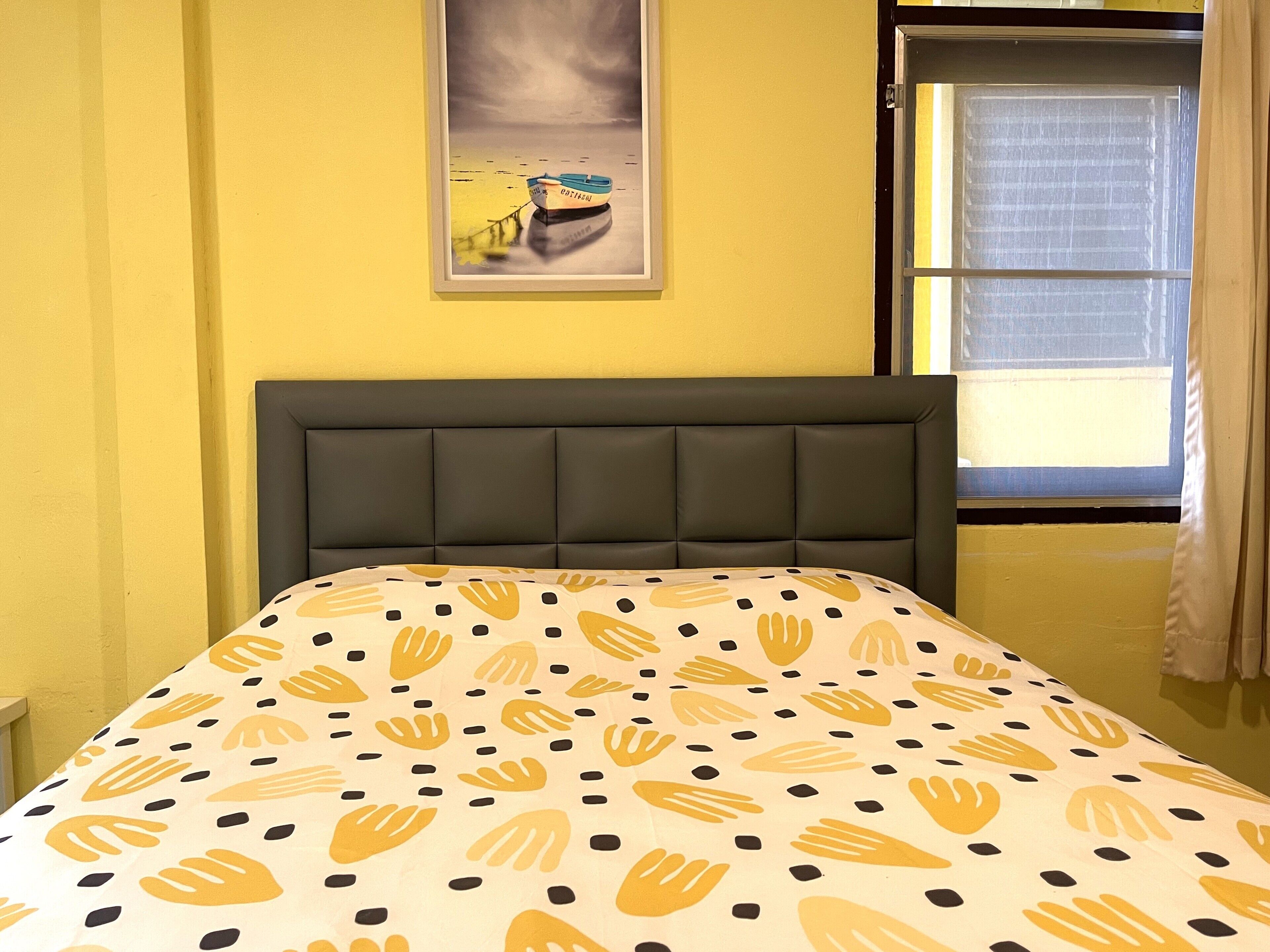 A bed with a patterned bedspread, a headboard, a framed picture on the wall, and a window with curtains.