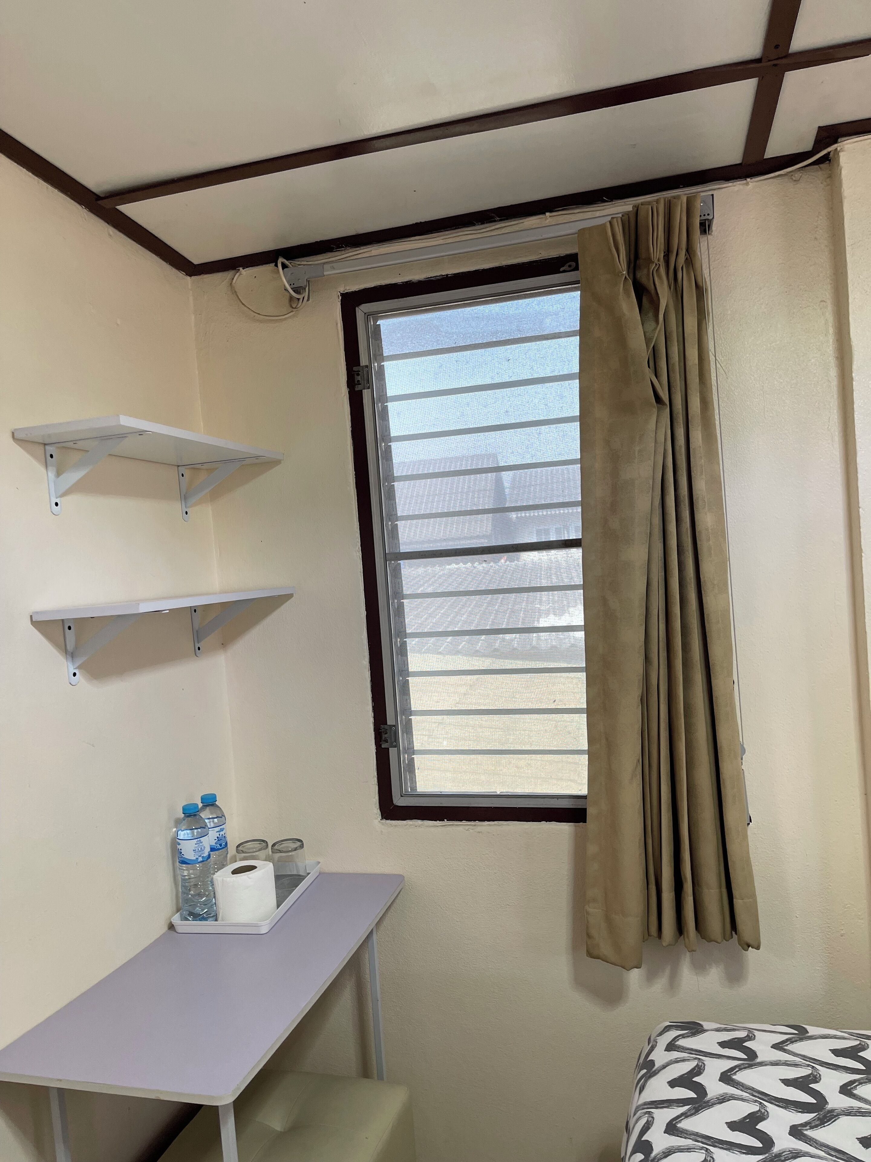 A room with a bed, a window with curtains, a desk with a chair, and two wall-mounted shelves.