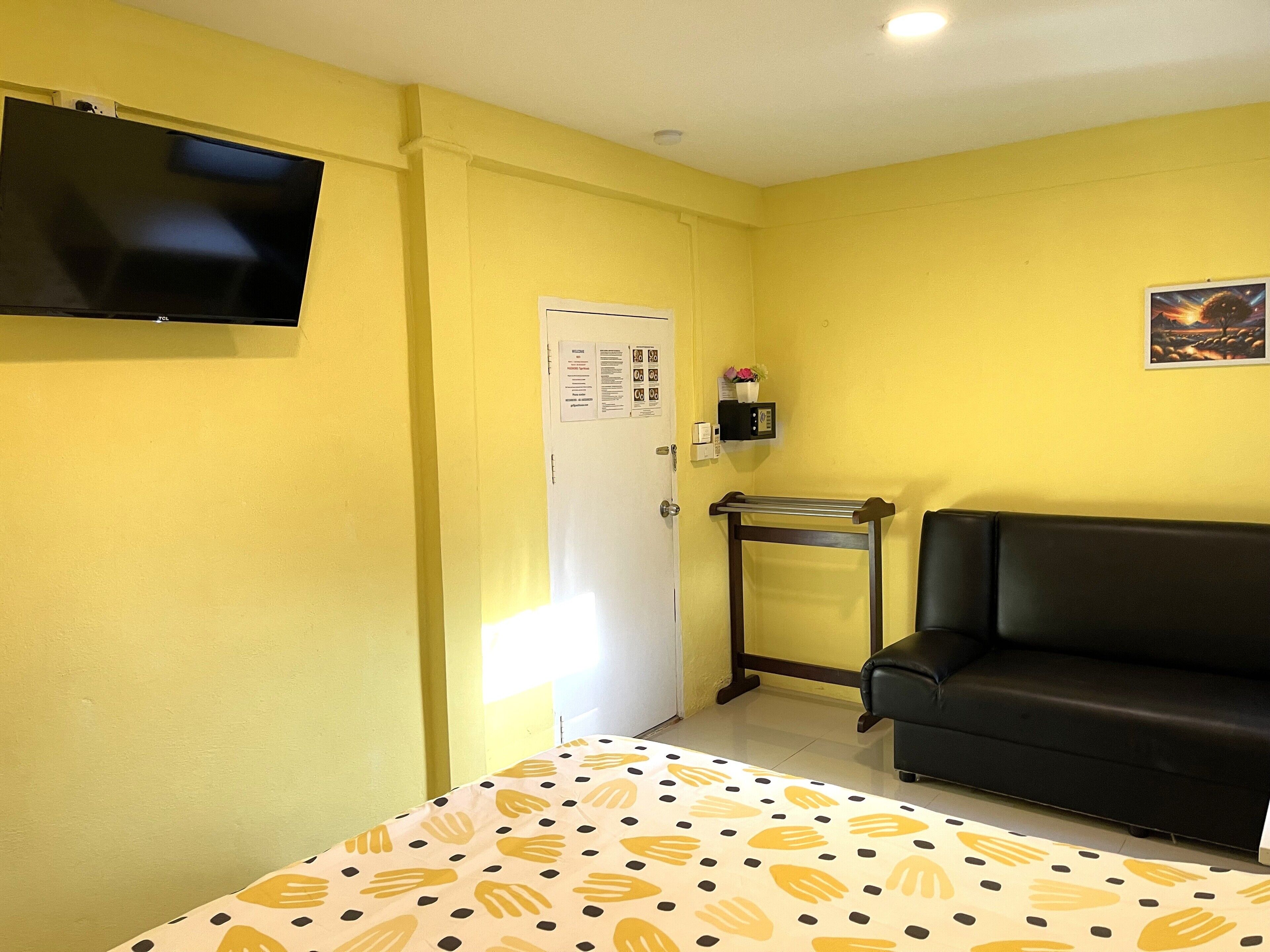 Una habitació amb un llit, un sofà negre, un televisor fixat a la paret, una porta amb un avís i una petita estant amb un vas.