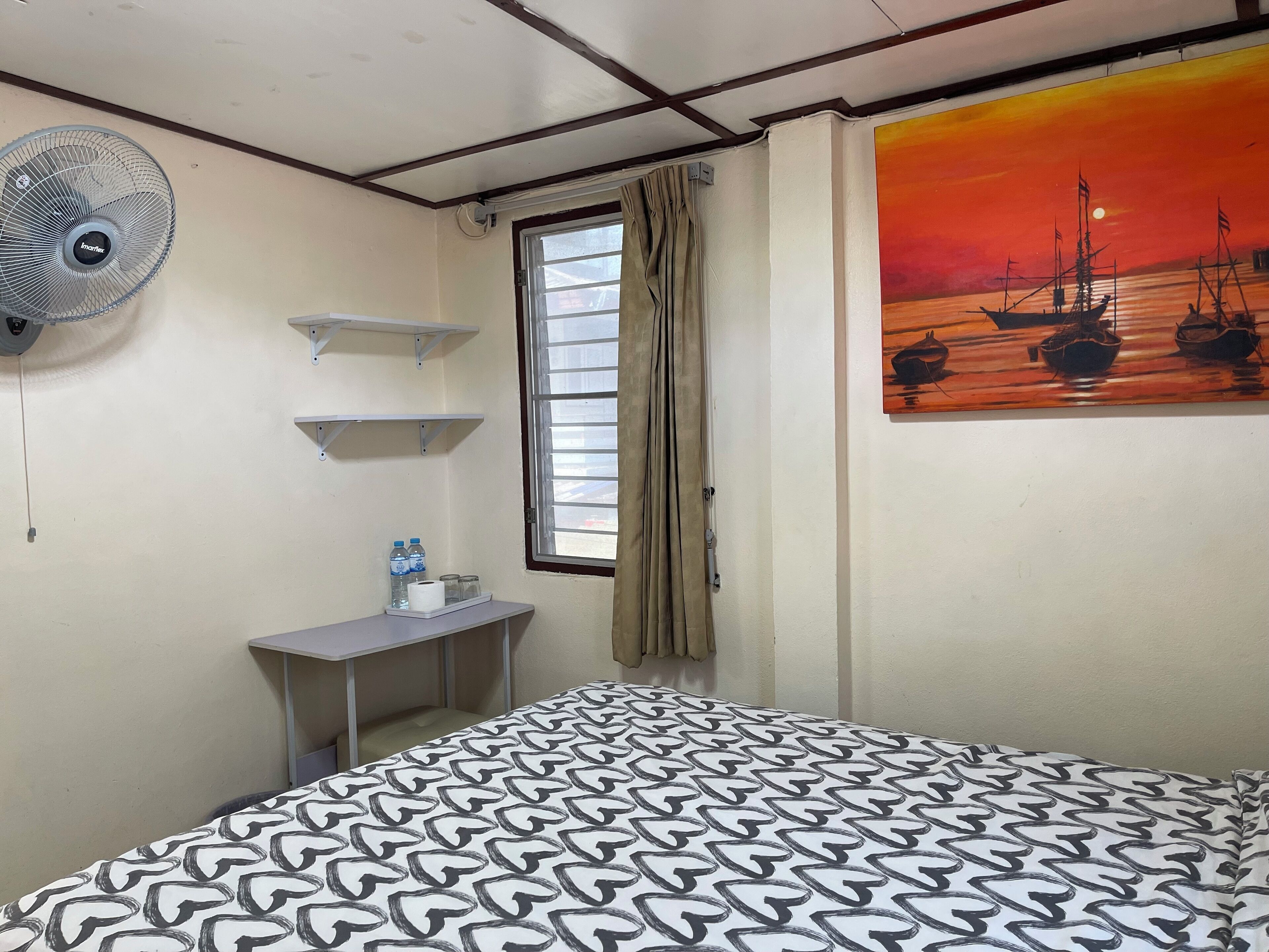 A bedroom with a patterned bedspread, a standing fan, a small desk with a water bottle, a painting of a sunset over water, and a window with curtains.