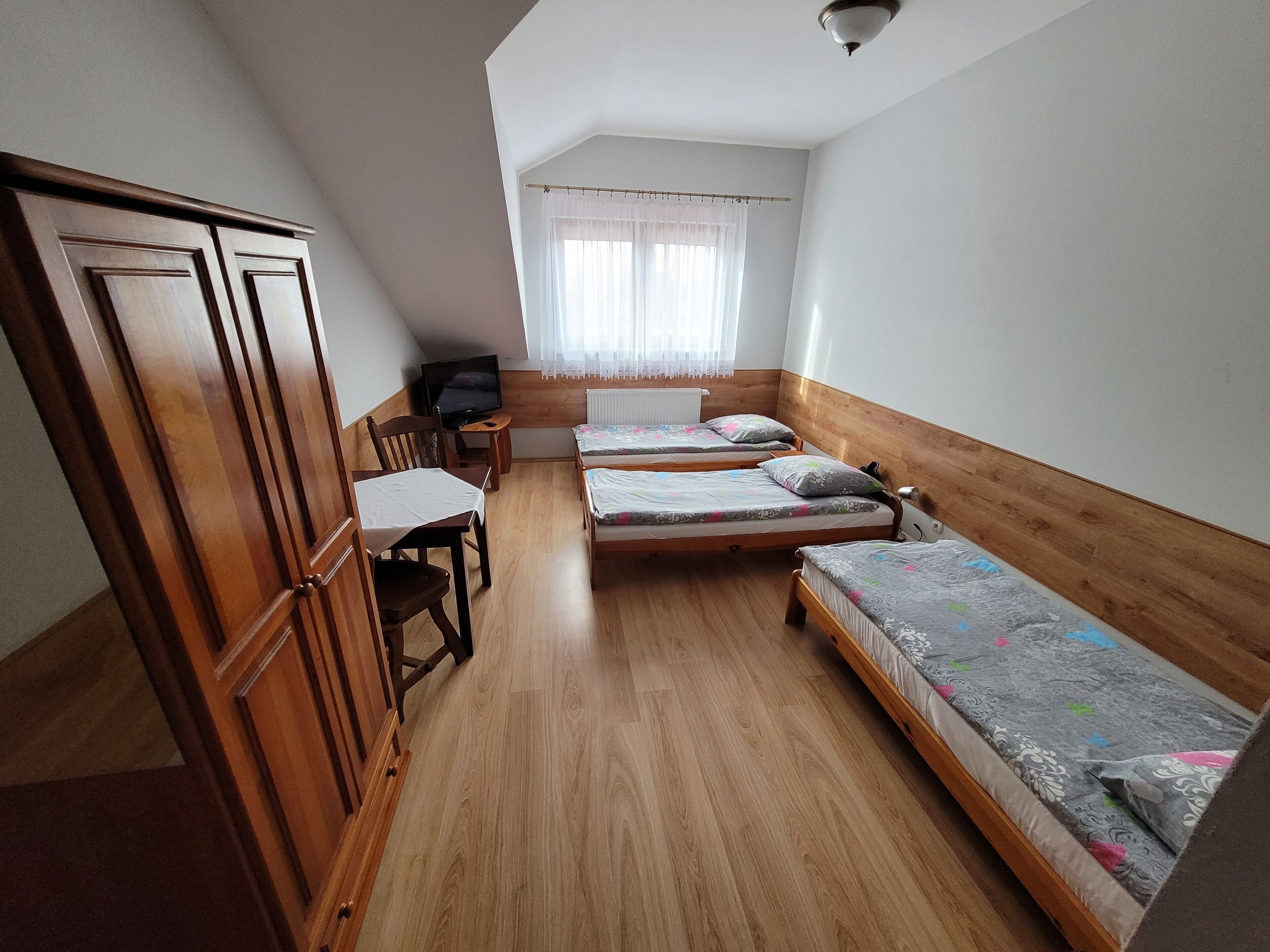 A small, cozy room with two single beds, a wooden wardrobe, a desk with a chair, and a television.