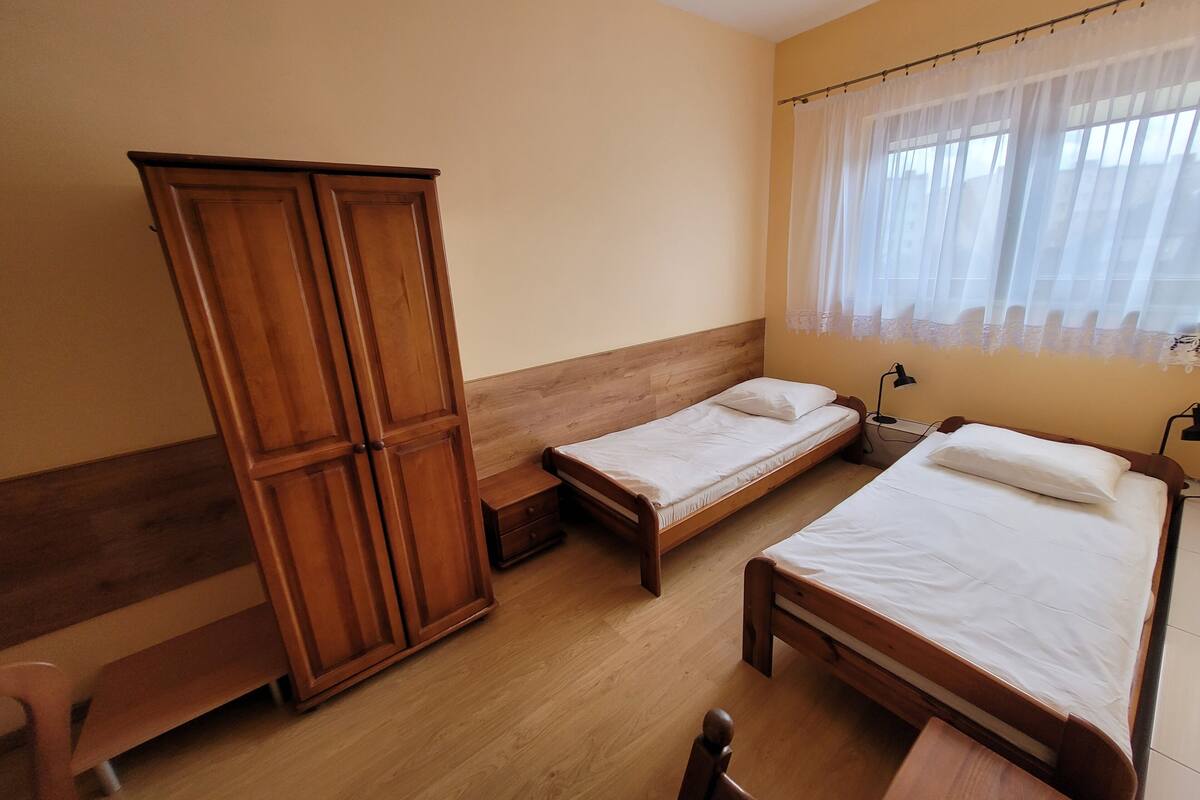 Une chambre avec deux lits simples, une armoire en bois, un banc et une fenêtre avec des rideaux légers.