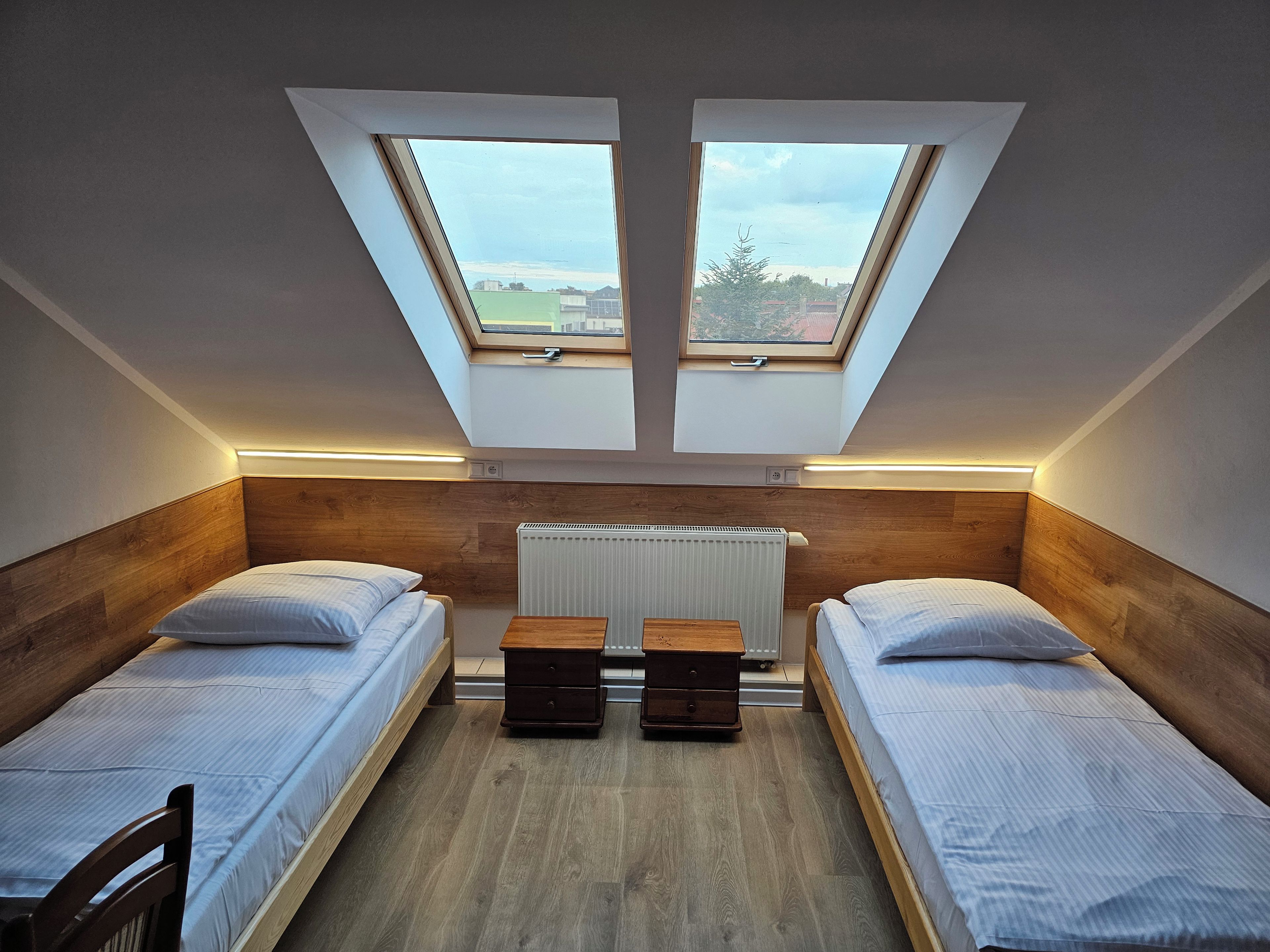 A small, modern room with two beds, a wooden headboard, a radiator, and a skylight.
