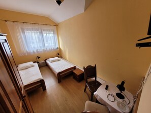 A small, cozy room with two single beds, a wooden table with a chair, and a window with sheer curtains.