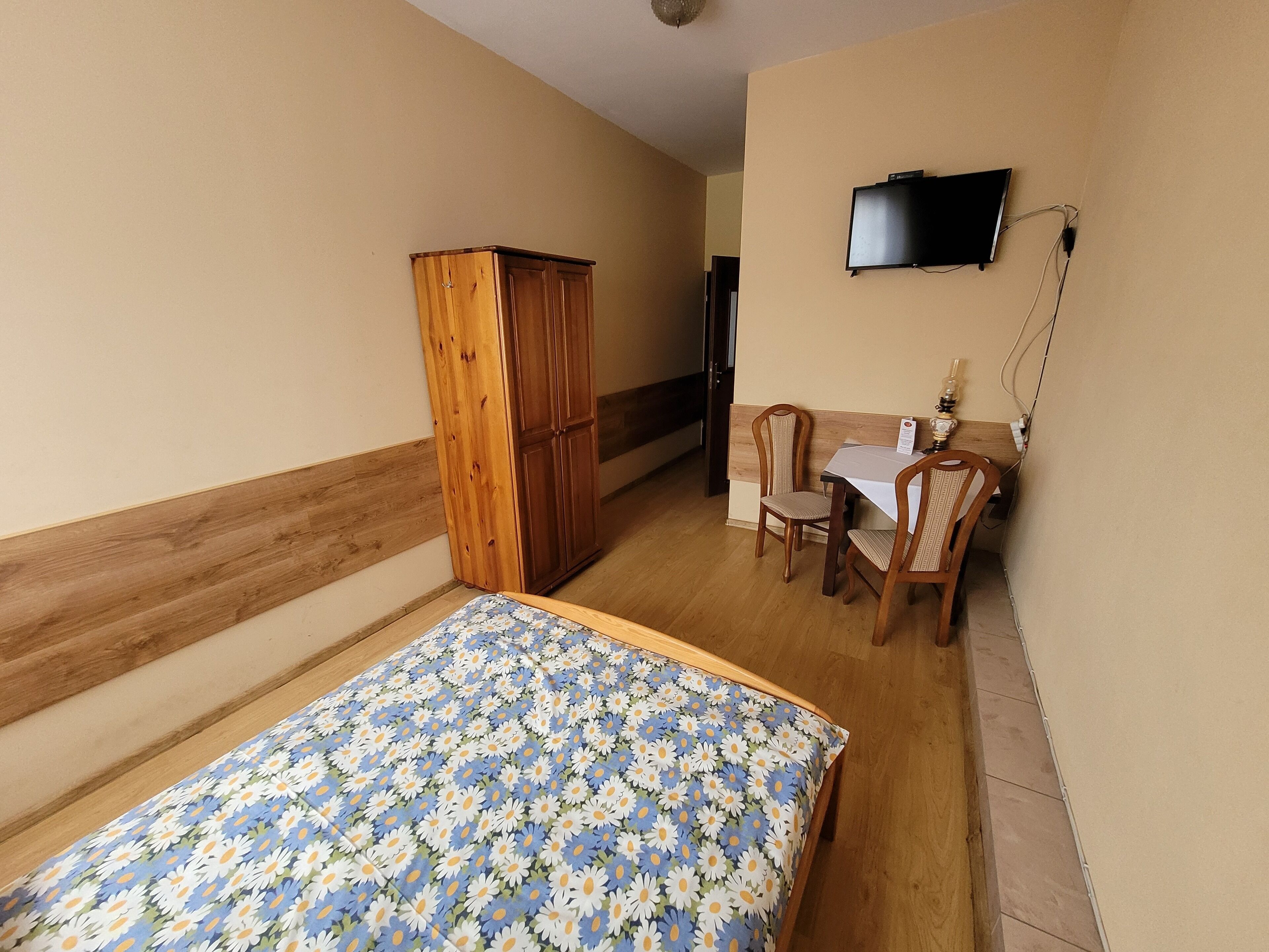 Een slaapkamer met een houten garderobe, een bed met een bloemenbedekking, een bureau met twee stoelen, en een televisie die aan de muur is bevestigd.