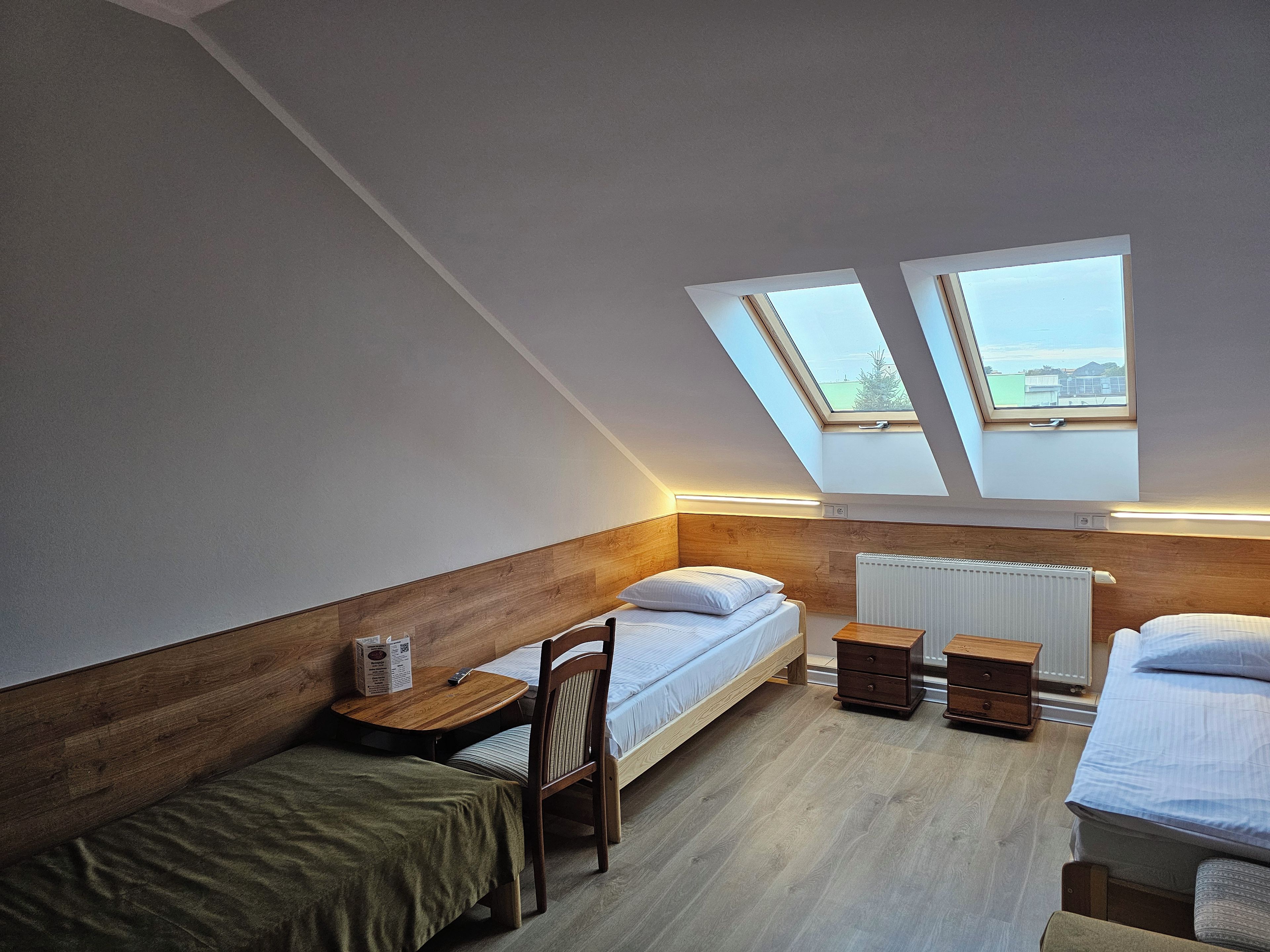 A small, modern bedroom with a sloped ceiling, two skylights, a wooden headboard, a single bed, a small table, and a chair.
