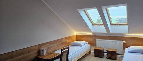 A small, modern bedroom with a sloped ceiling, two skylights, a wooden headboard, a single bed, a small table, and a chair.