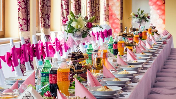 Una larga mesa de banquete dispuesta con platos, vasos y botellas, adornada con globos y cintas rosas y blancas.