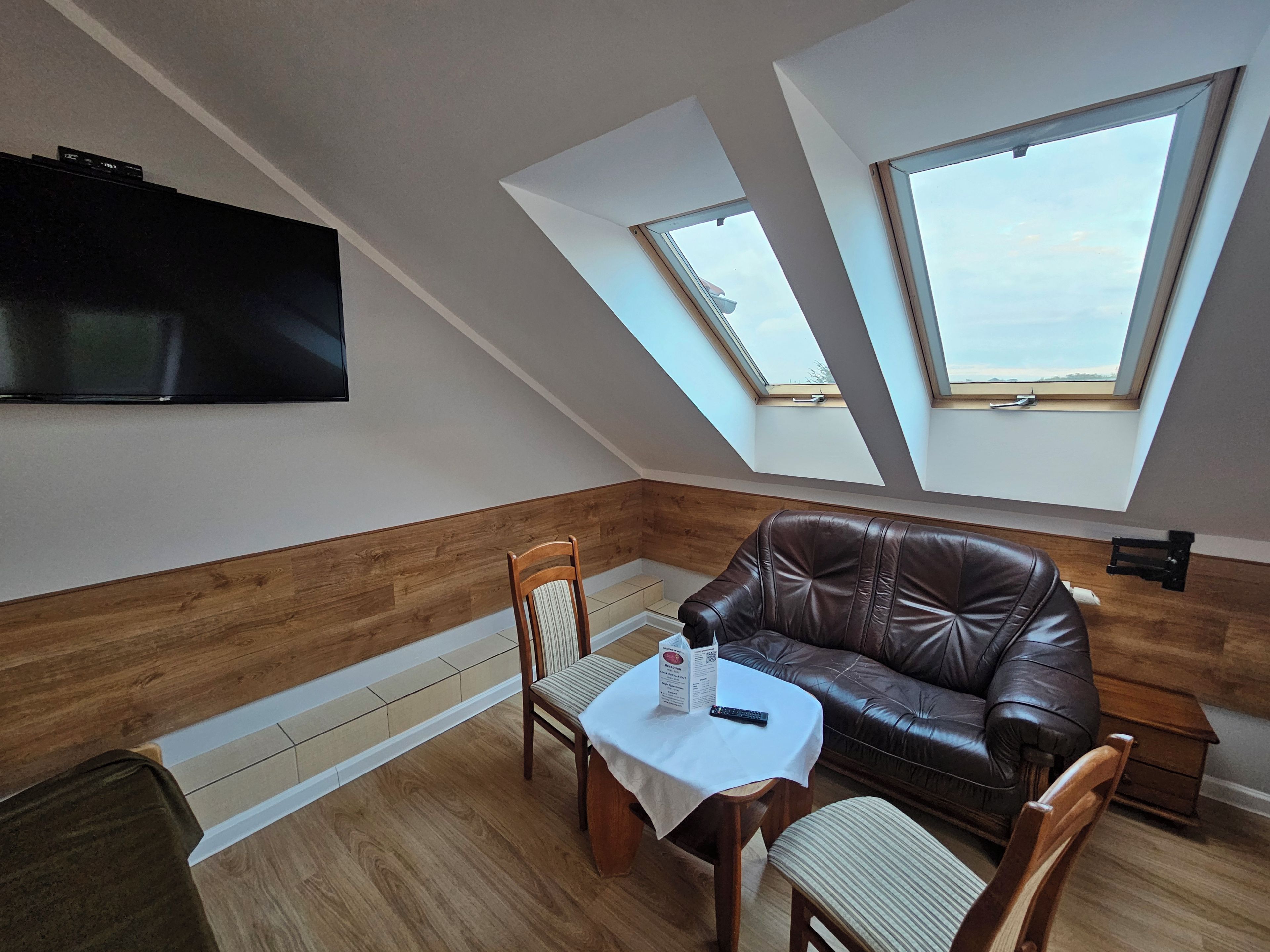 Una habitación con sofá de cuero marrón, una mesa de comedor de madera con sillas, televisor de pantalla plana en la pared y tragaluces que dejan entrar luz natural.