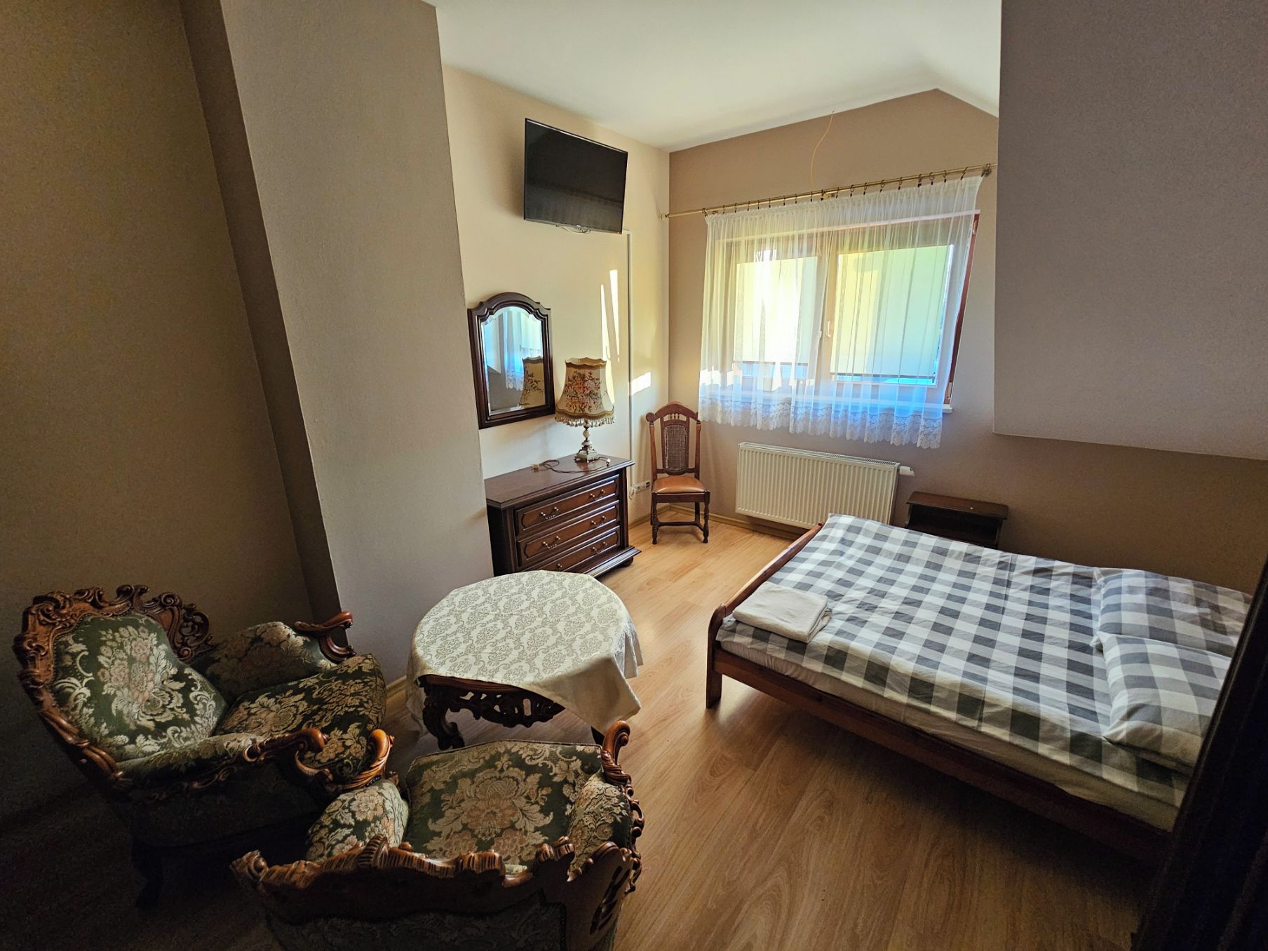A bedroom with a bed, a chair, a small table, a dresser, a mirror, a radiator, and a window with curtains.