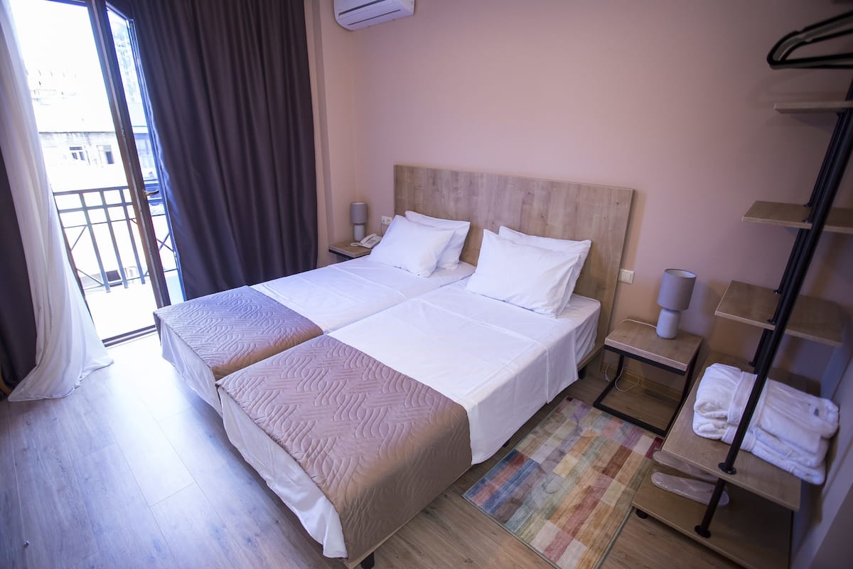 A double bed with a wooden headboard, a bedside table with a lamp, a small shelf with towels, and a view of the outside through a window with curtains.