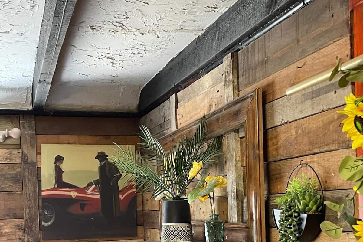 A rustic room with wooden walls, a bed with patterned bedding, a shelf with plants, and a framed picture on the wall.