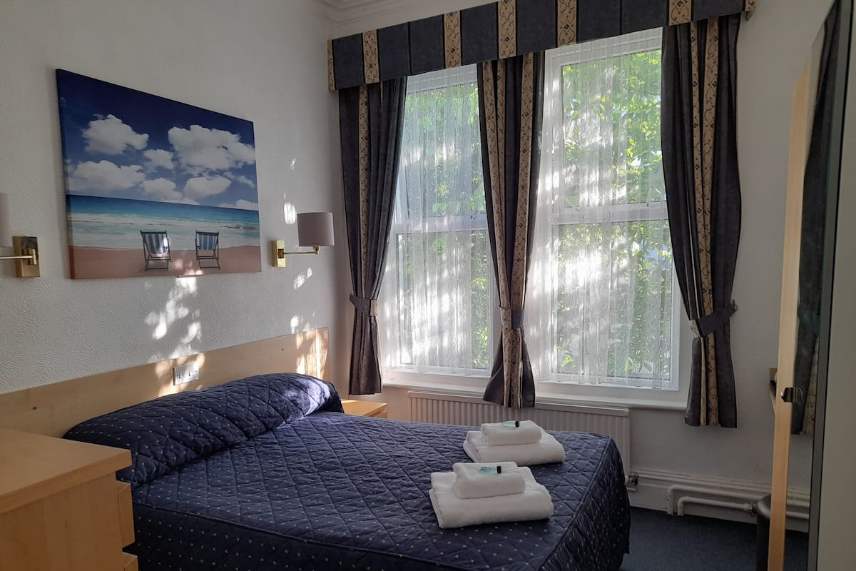 A hotel room with a bed, a painting of a beach scene, a window with curtains, and a view of greenery outside.