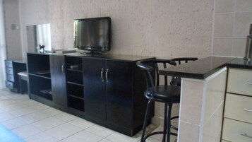 Una cocina con encimera negra, televisor sobre el lavabo y un taburete de bar.